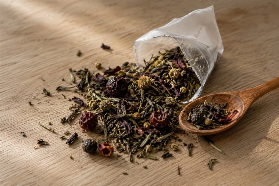 Loose dried herbal tea leaves, chamomile flowers and dried berries spilling from an open pyramid tea bag onto a wooden table, with a wooden spoon beside them
